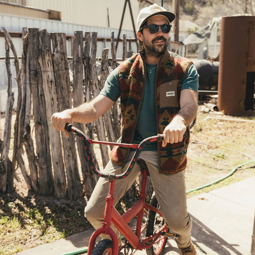 Man with a beard wearing sunglasses and a patterned vest standing next to a red bicycle outdoors.