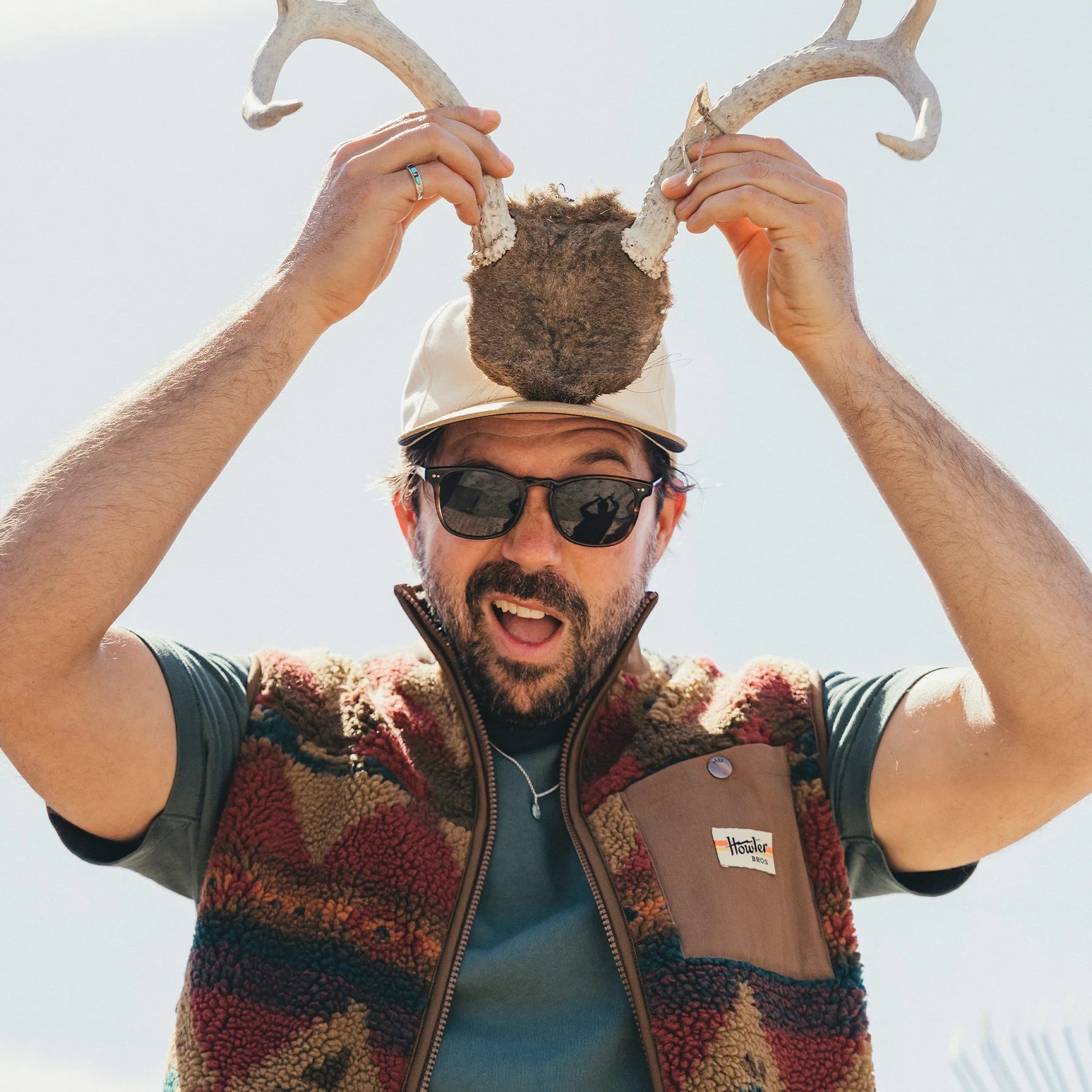 Man wearing a cap with antlers and sunglasses, holding antlers above his head against a clear sky.