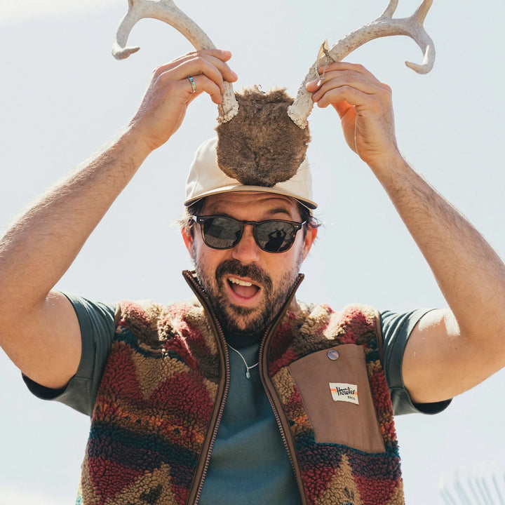 Man wearing a cap with antlers and sunglasses, holding antlers above his head against a clear sky.
