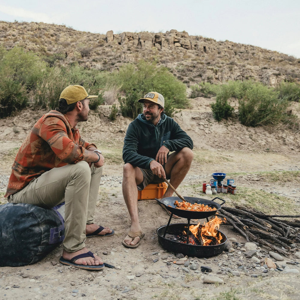Two men sitting by a campfire in a desert setting, cooking food on a grill.