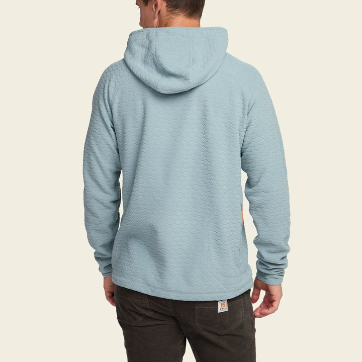 Person wearing a light blue hoodie with a visible brand logo on the chest, standing against a beige background.