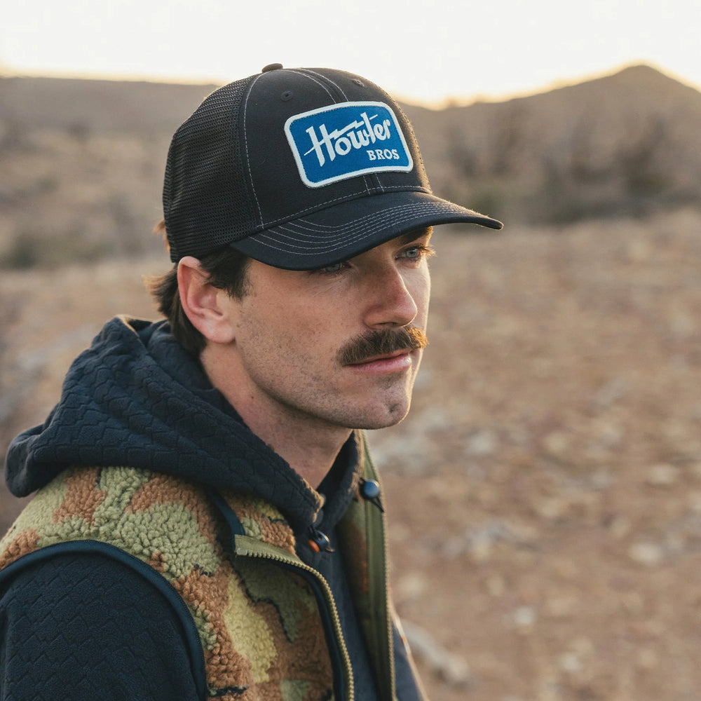 Man wearing a black cap with a logo, standing in a desert-like landscape.
