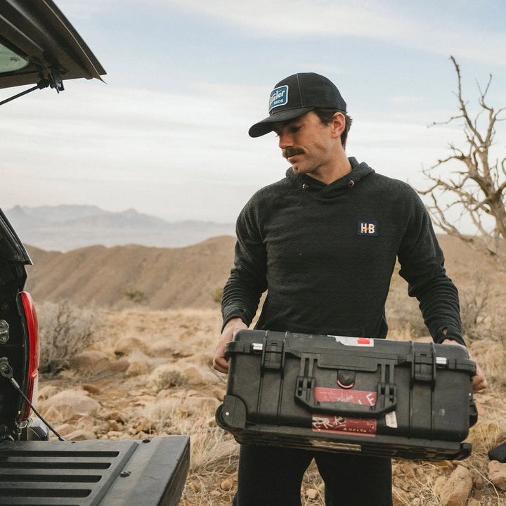 Man holding a black toolbox in a desert setting with mountains in the background