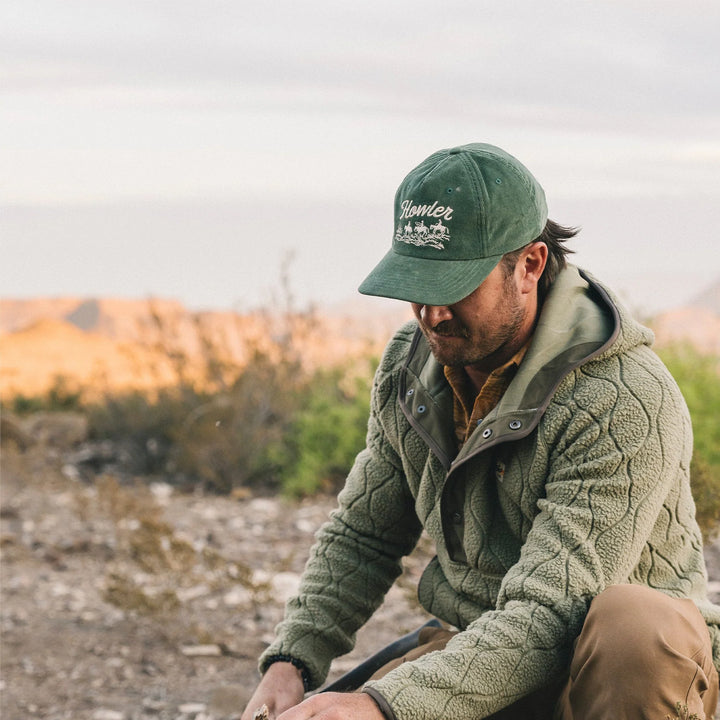 Man wearing a green cap and jacket sitting outdoors with a blurred natural background