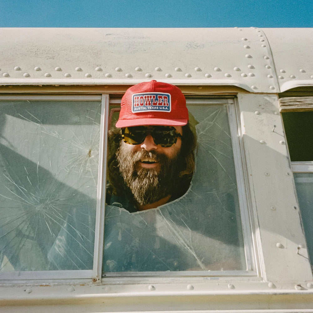 Person wearing a red cap with 'Howler' branding, peeking out of a window with a cracked glass panel.