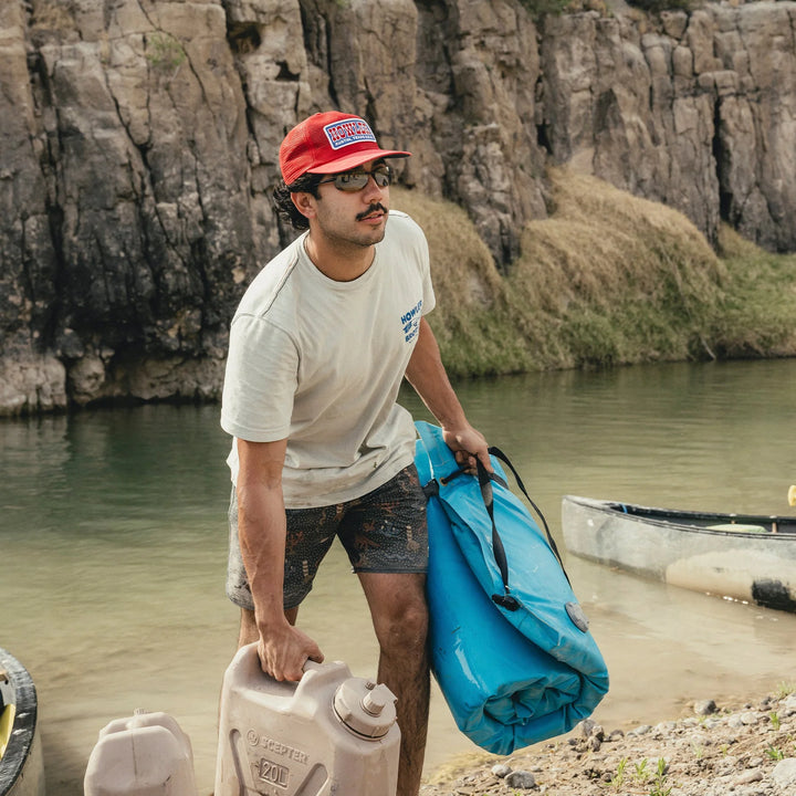 Man with a red cap and sunglasses holding a blue bag and a water container by a river.