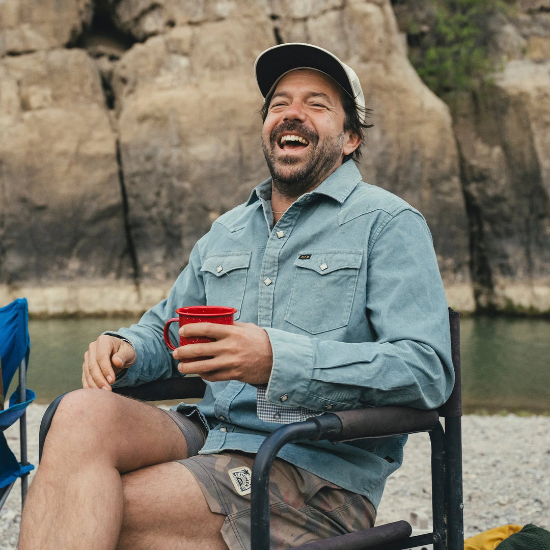 Man sitting outdoors by a body of water, holding a red mug and smiling.