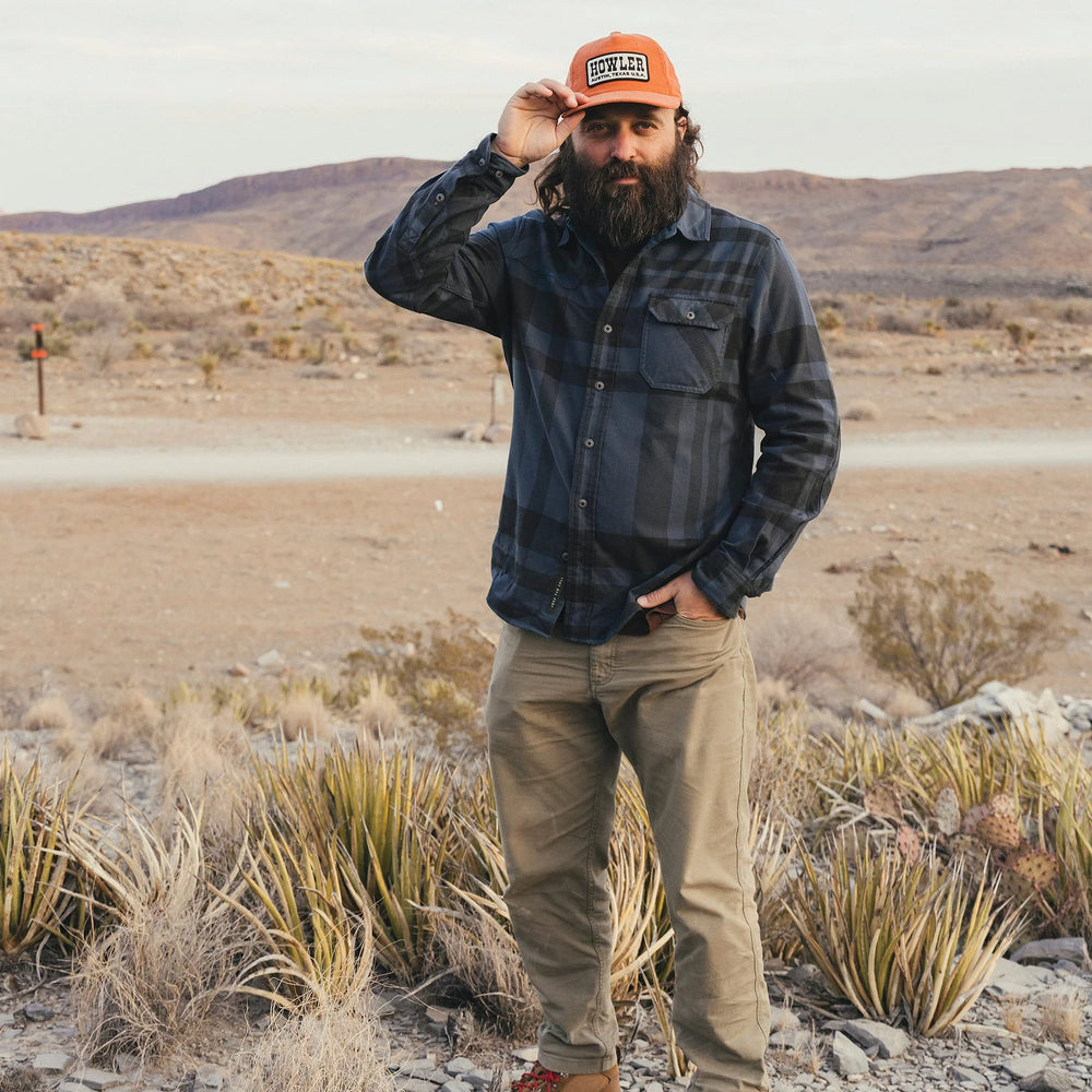 Man standing in a desert landscape wearing a plaid shirt and cap.