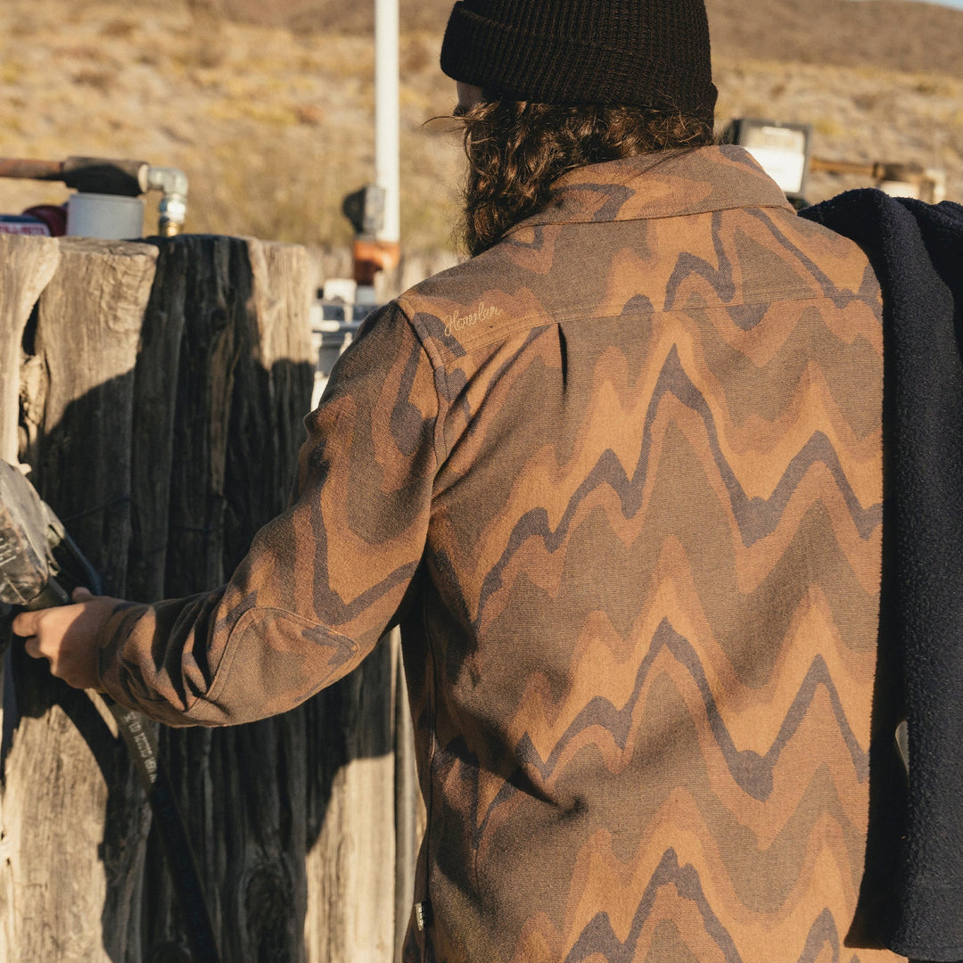 Person wearing a brown patterned jacket standing next to a wooden fence outdoors.