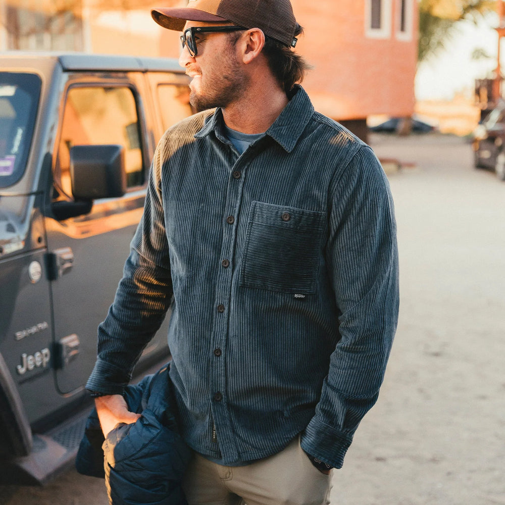Man in a blue corduroy shirt and brown cap standing next to a Jeep.