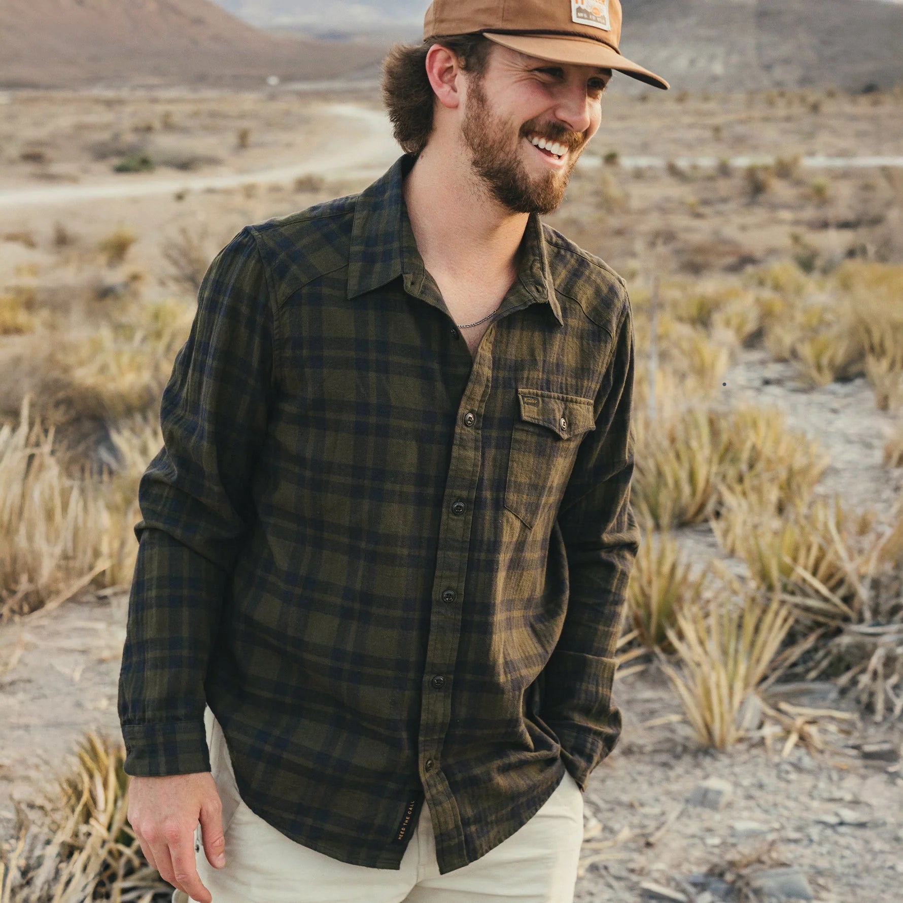 Man wearing a Howler Brothers plaid shirt and cap standing in a desert landscape