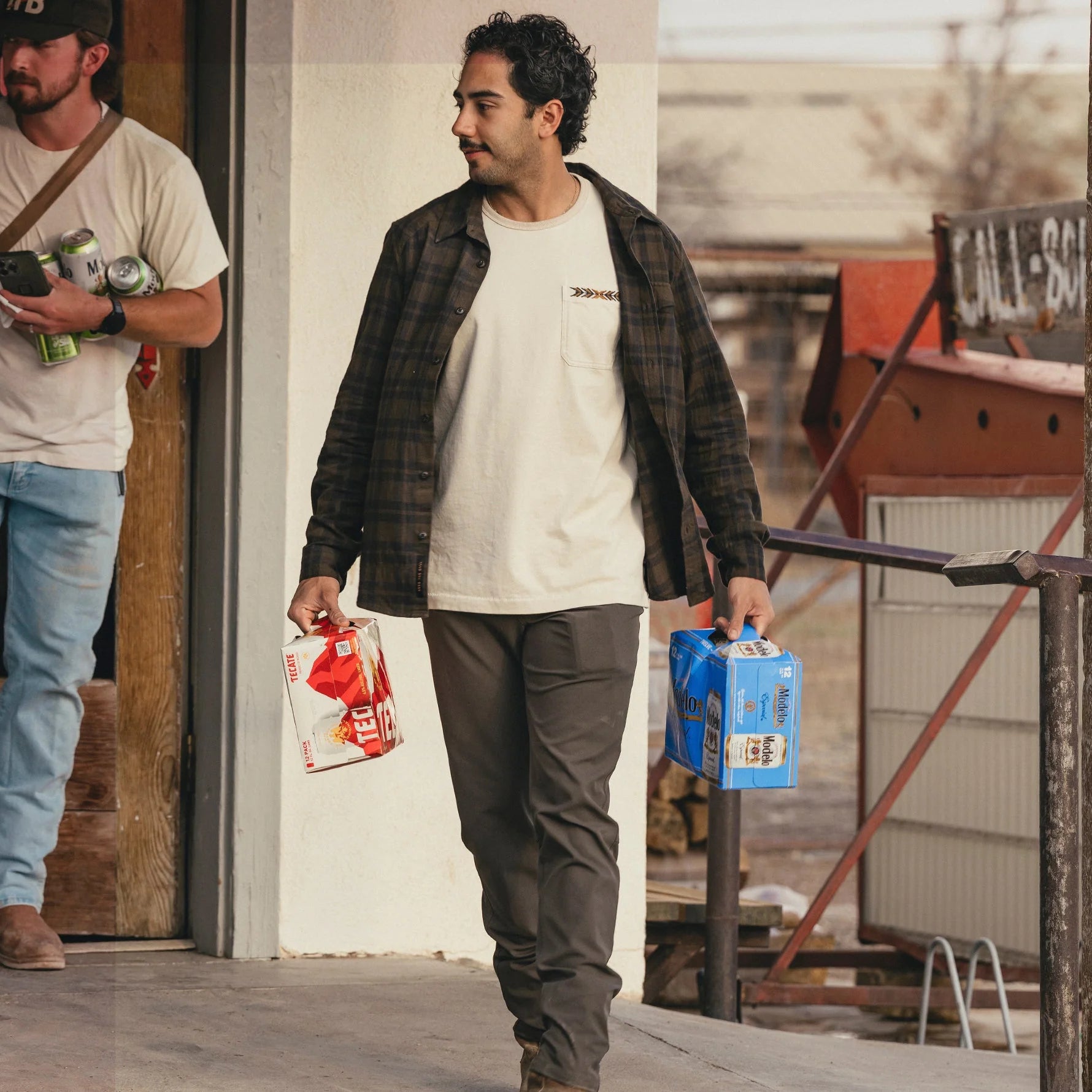 Man walking with a six-pack of beer and a case of pop, standing in front of a white wall, wearing a Howler Brothers plaid shirt
