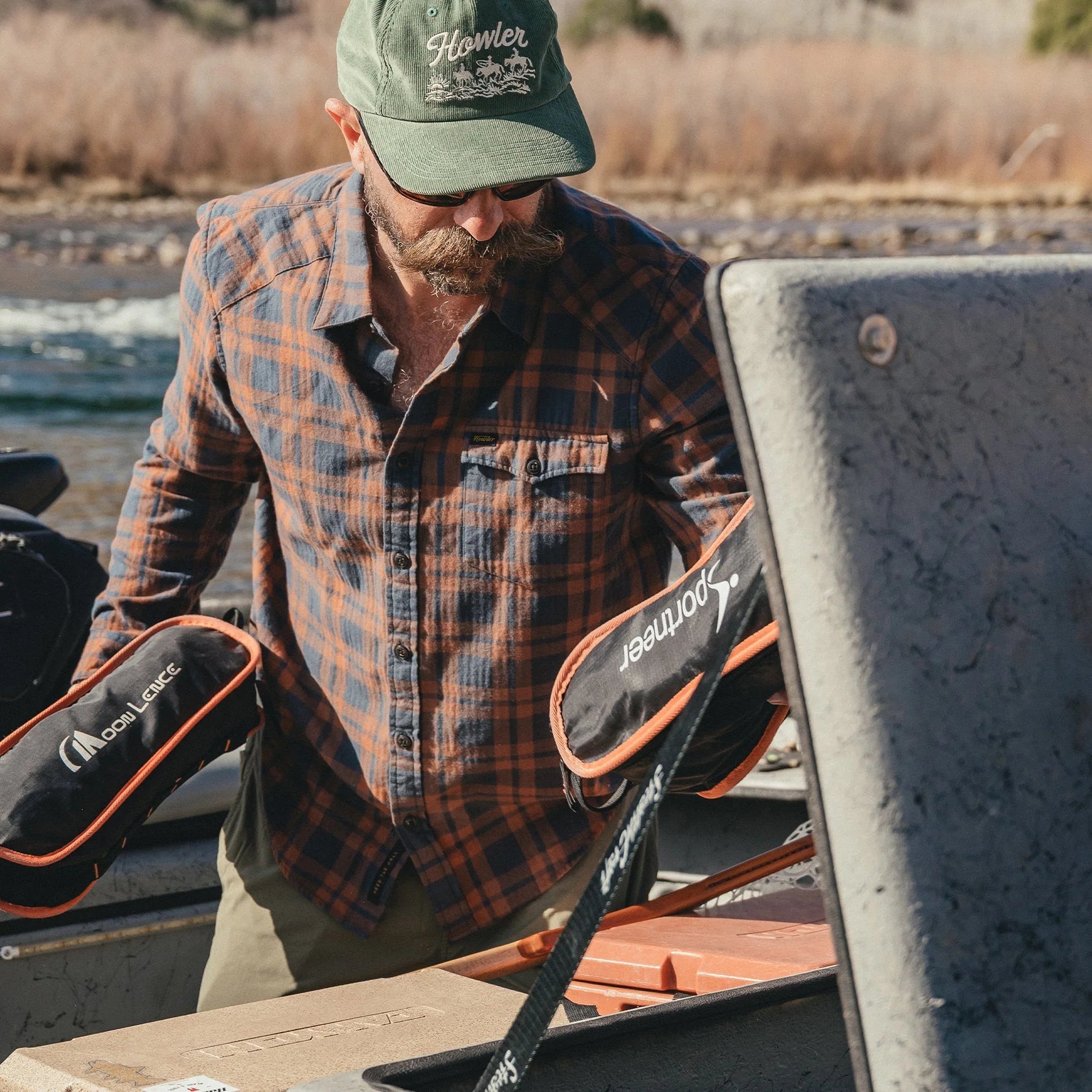 Man in Howler Brothers plaid shirt and cap with outdoor gear by a body of water