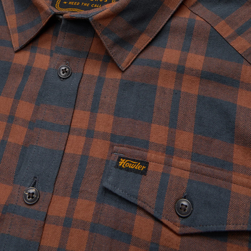 Close-up of a Howler Brothers plaid shirt with a brand label on a brown and blue checkered pattern.