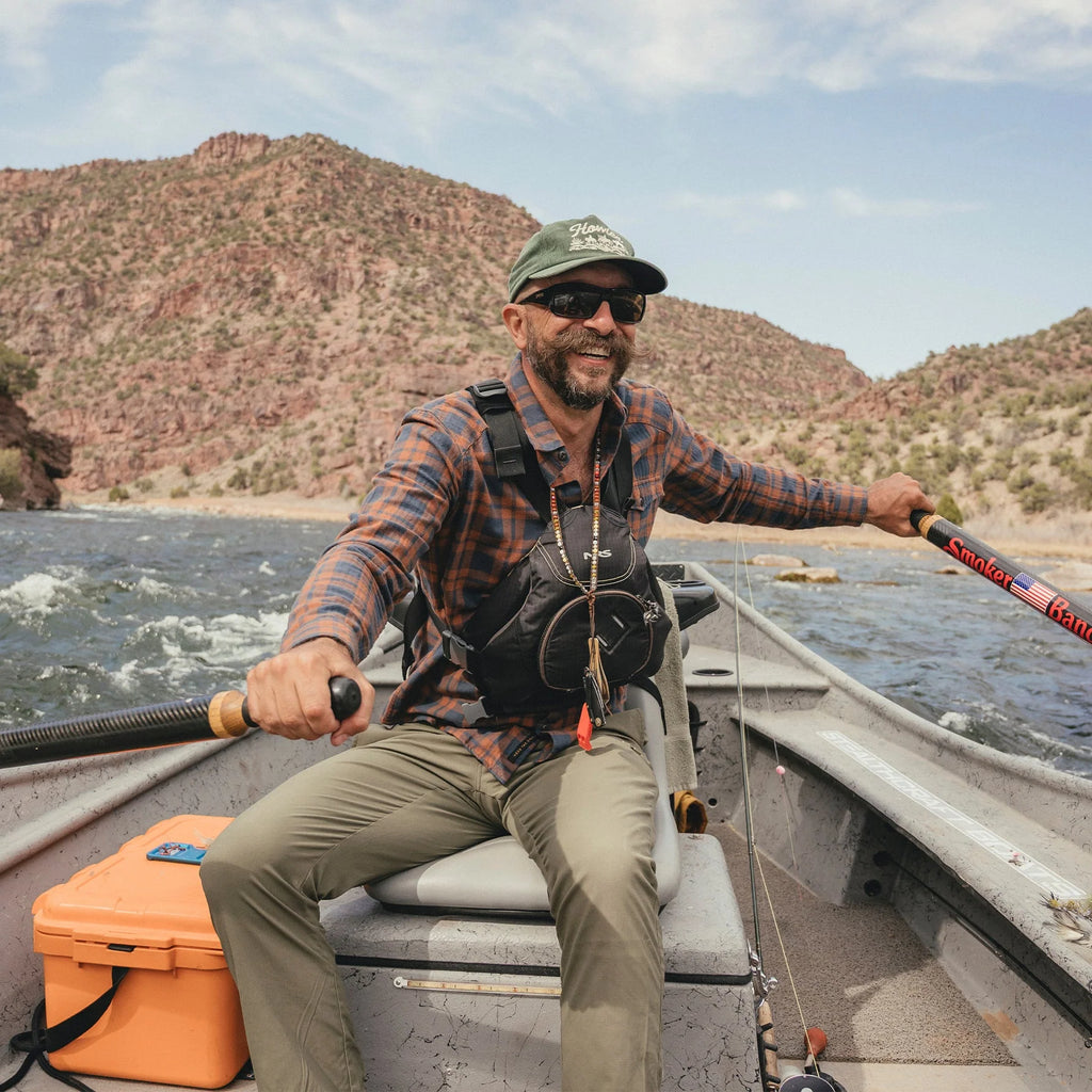 Man fishing on a boat with mountains in the background, wearing a Howler Brothers plaid shirt