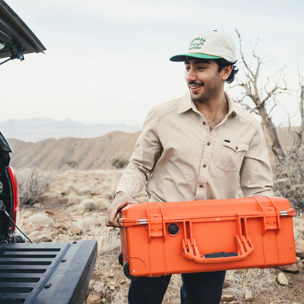 Man holding an orange case in a desert setting with mountains in the background, wearing a flannel