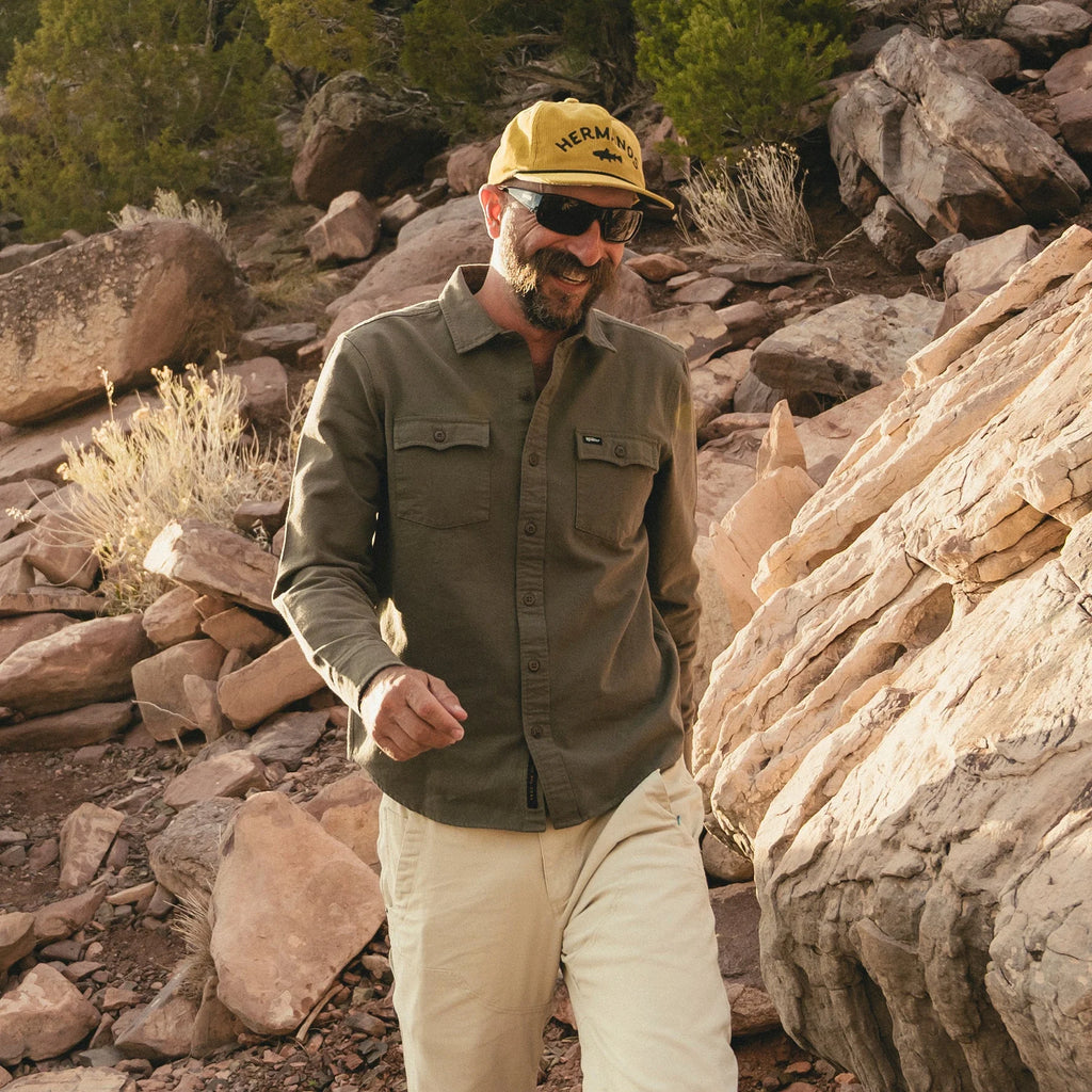 Man wearing a green shirt, beige pants, and a yellow cap with sunglasses, standing in a rocky desert landscape.