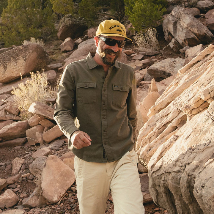 Man wearing a green shirt, beige pants, and a yellow cap with sunglasses, standing in a rocky desert landscape.