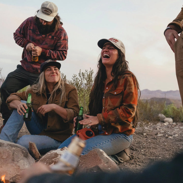 Three people sitting around a campfire in a desert setting, enjoying drinks.