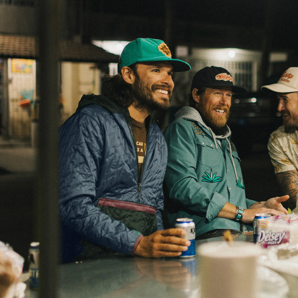 Three men sitting together, smiling and enjoying drinks at a table, wearing Howler Brothers Voltage Quilted Pullover