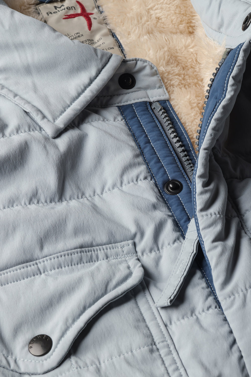 Close-up of a light blue puffer jacket with a beige sherpa lining, featuring a blue zipper, snap buttons, and a collar.