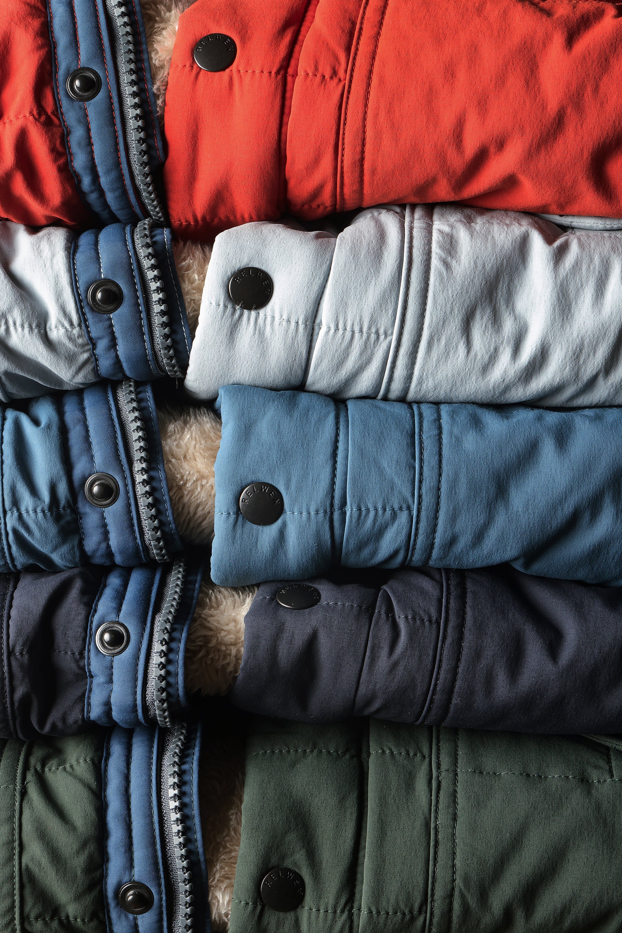 A close-up stack of folded quilted winter jackets in multiple colors, including red, light blue, medium blue, navy, and olive green, with visible snap buttons and a fuzzy lining.