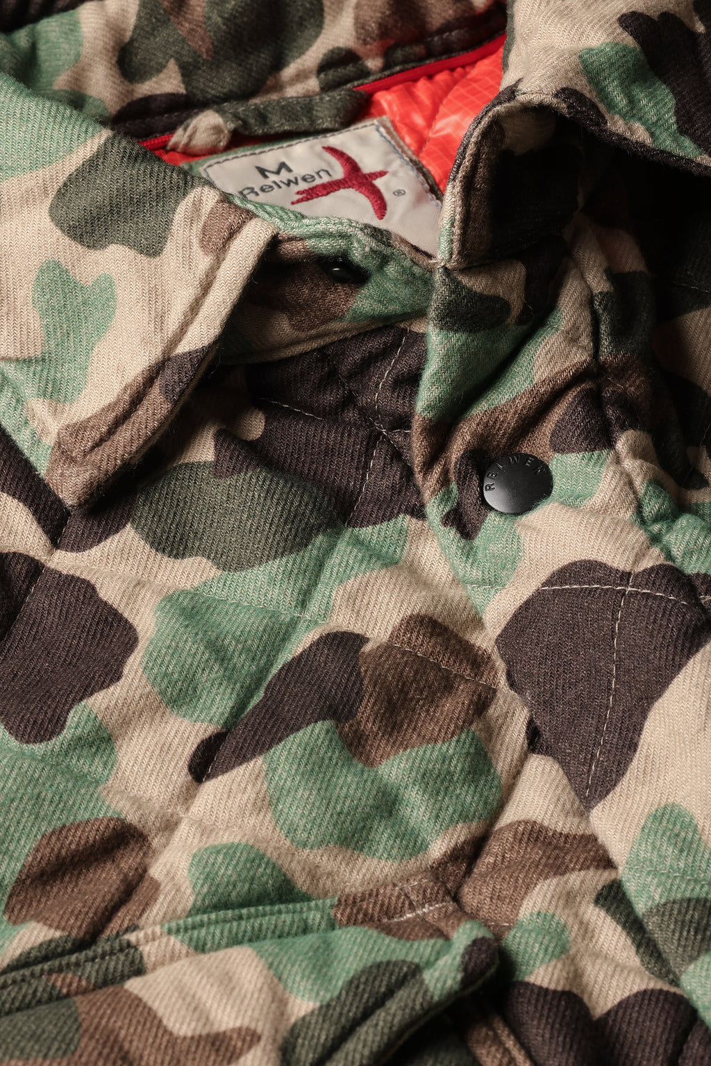 Close-up of a camouflage jacket with a green, brown, and beige camo pattern, featuring a visible neck label for 'M Reifken'.