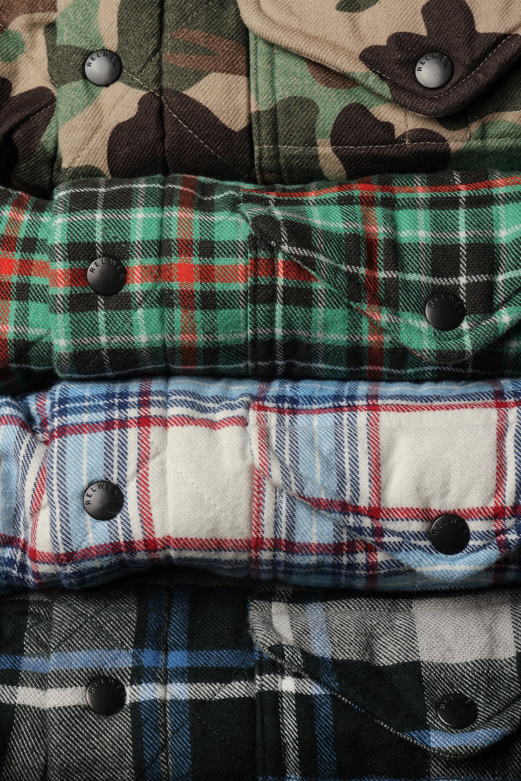 A close-up stacked arrangement of folded plaid and camouflage flannel shirts, showing snap-button closures and fabric textures in green, blue, red, black, and tan tones.