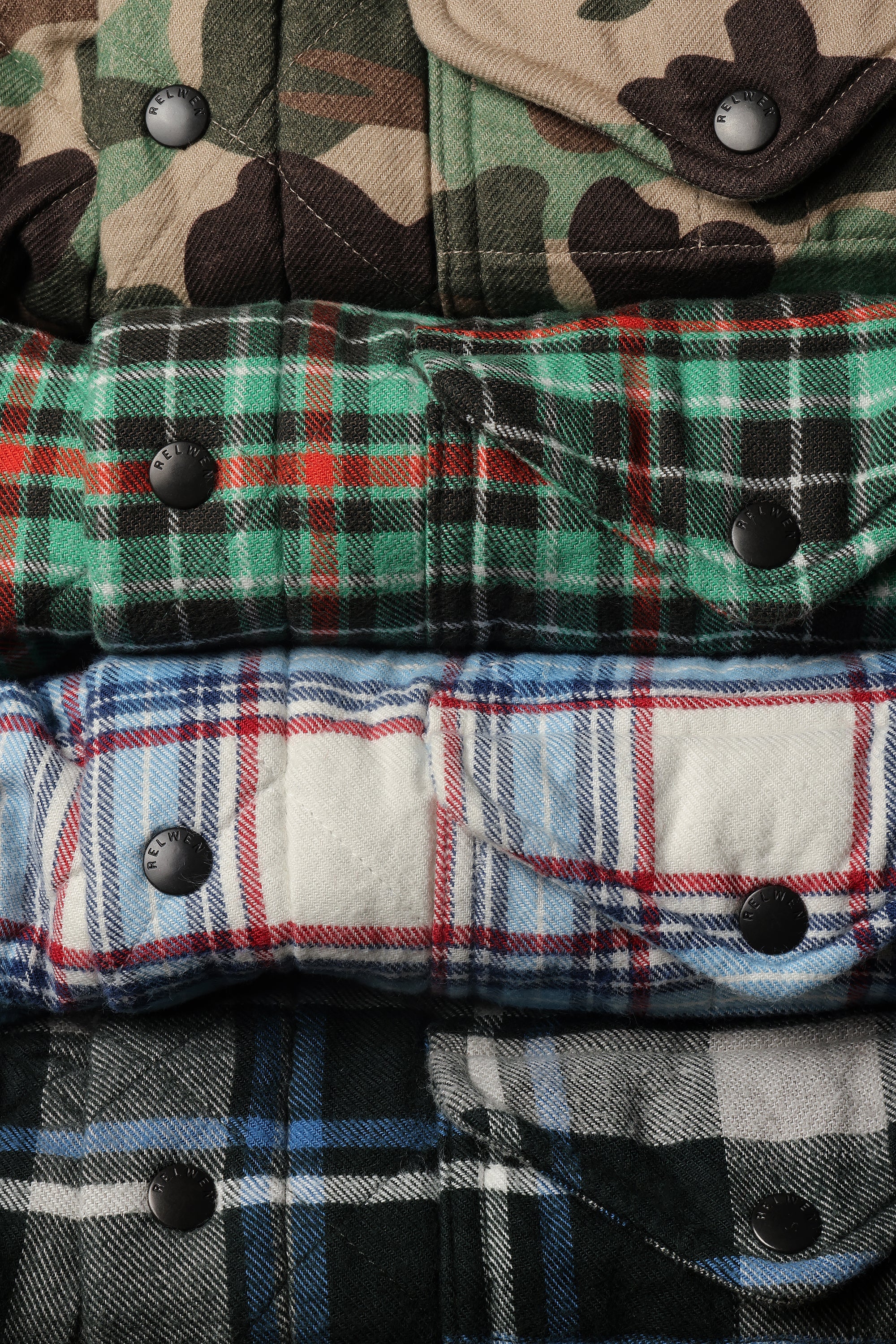 A close-up stacked arrangement of folded plaid and camouflage flannel shirts, showing snap-button closures and fabric textures in green, blue, red, black, and tan tones.