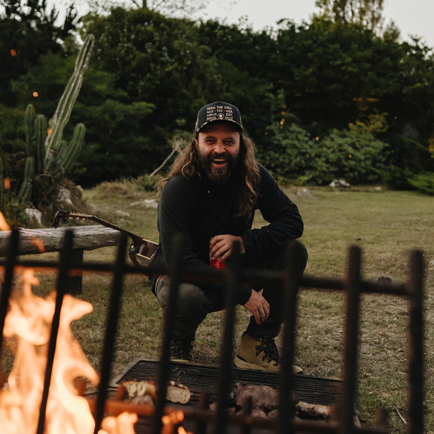 A smiling man with long hair and a beard crouches in front of a campfire outdoors, wearing a black hoodie, dark pants, tan boots, and a black baseball cap with a gold logo. Trees and grass are visible in the background.