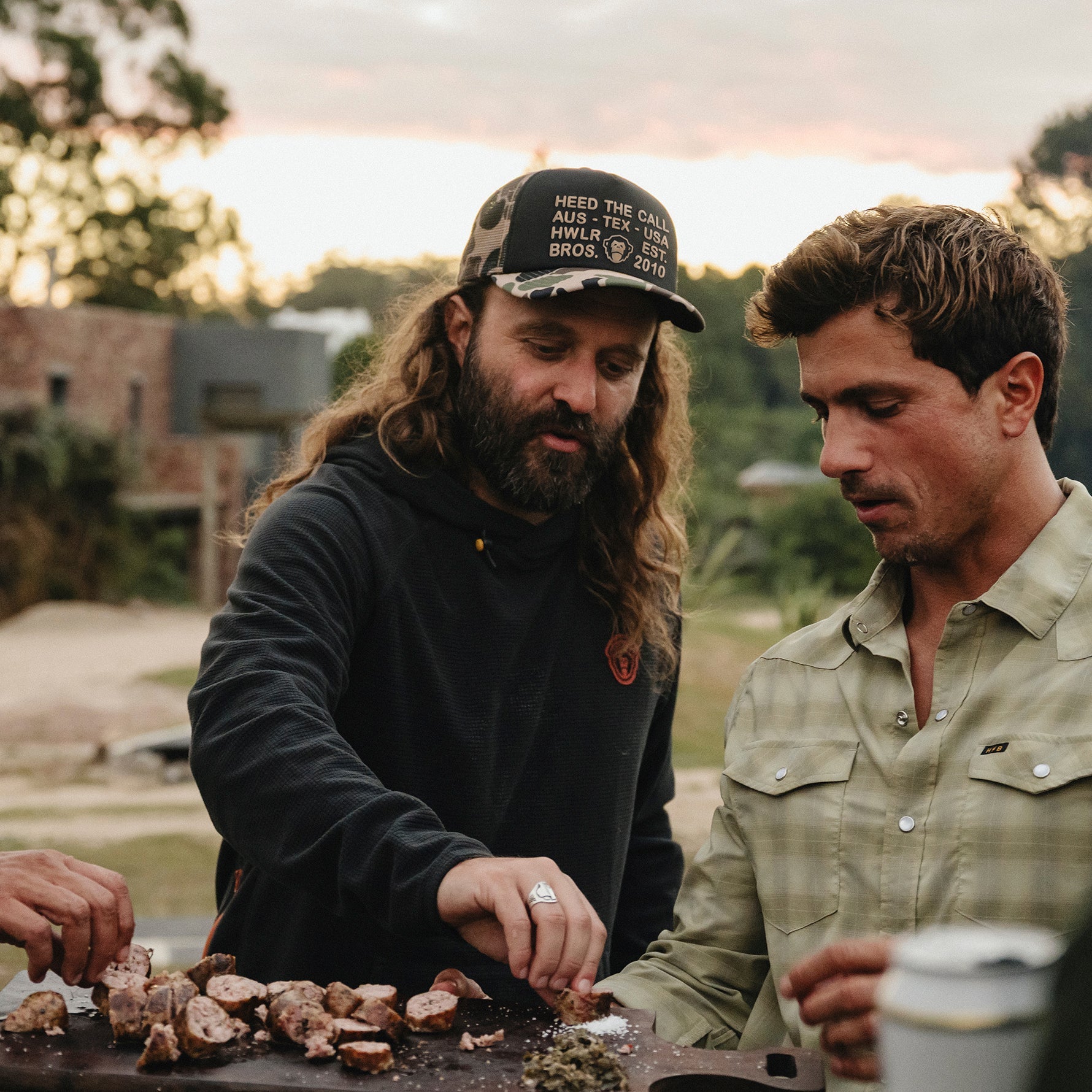 Two men standing outdoors at dusk near a grill, with one man wearing a black hoodie and a camouflage baseball cap with white text, and the other wearing a light olive green button-up shirt.