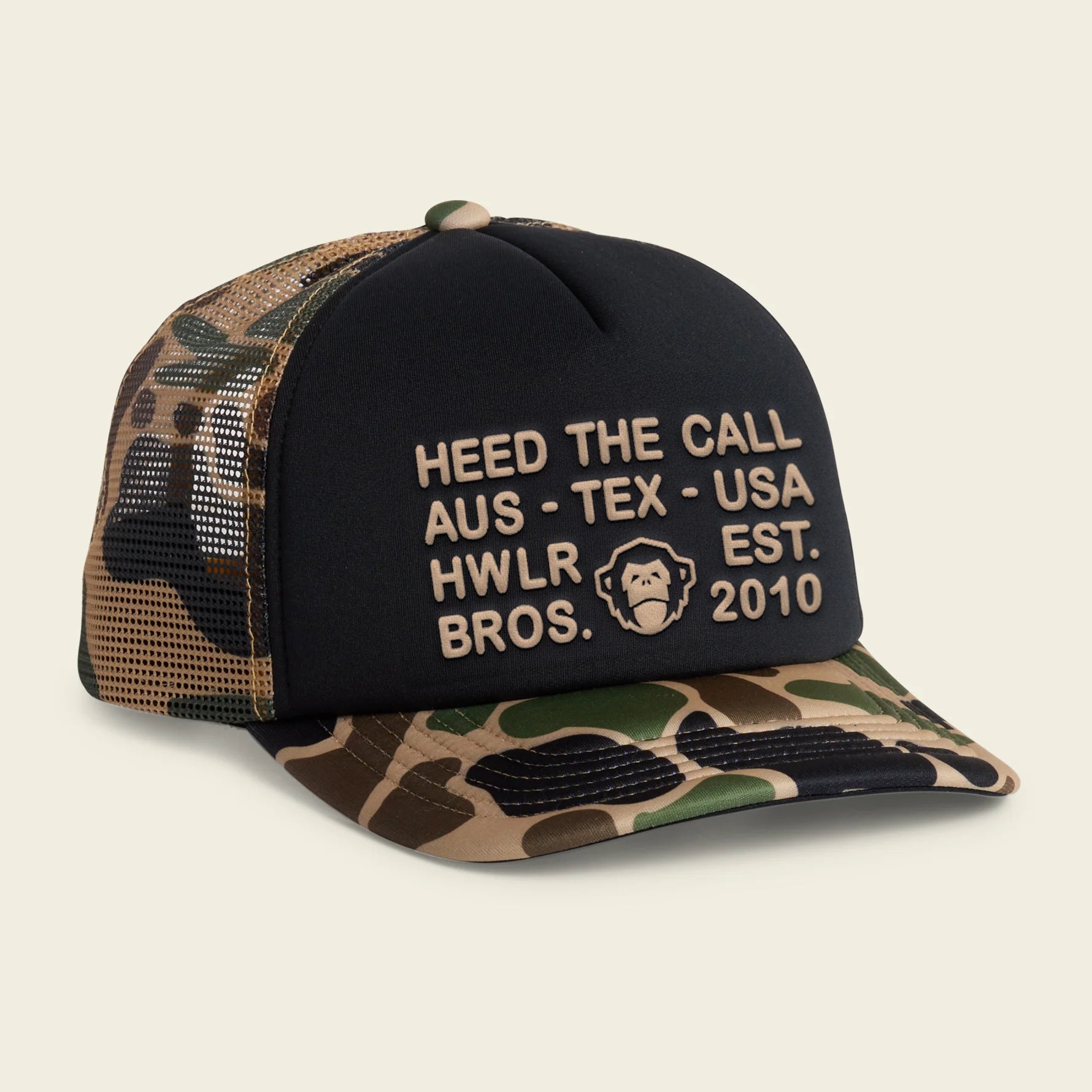 A trucker-style cap with a black front panel and camouflage-patterned mesh back and brim. The front panel features beige embroidered text reading 'HEED THE CALL AUS - TEX - USA HWLR EST.