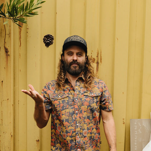A man with a beard and long curly hair stands in front of a yellow corrugated metal wall, wearing a black baseball cap with white text and a short-sleeve button-up shirt with a colorful floral and botanical pattern. He is gesturing with his right hand, and a pinecone floats near his head.