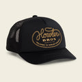 A black trucker hat with a curved brim and black mesh back panels, featuring a gold embroidered logo on the front that reads 'Howler Bros. Austin, TX' with 'Welcome to Howler County' below, set against a light beige background.