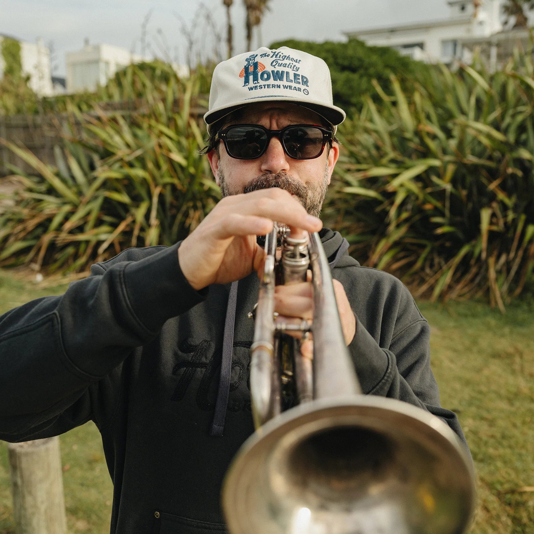 A man wearing a white 'Howler Western Wear' cap, black sunglasses, and a black hoodie is playing a silver trumpet outdoors in front of green plants.