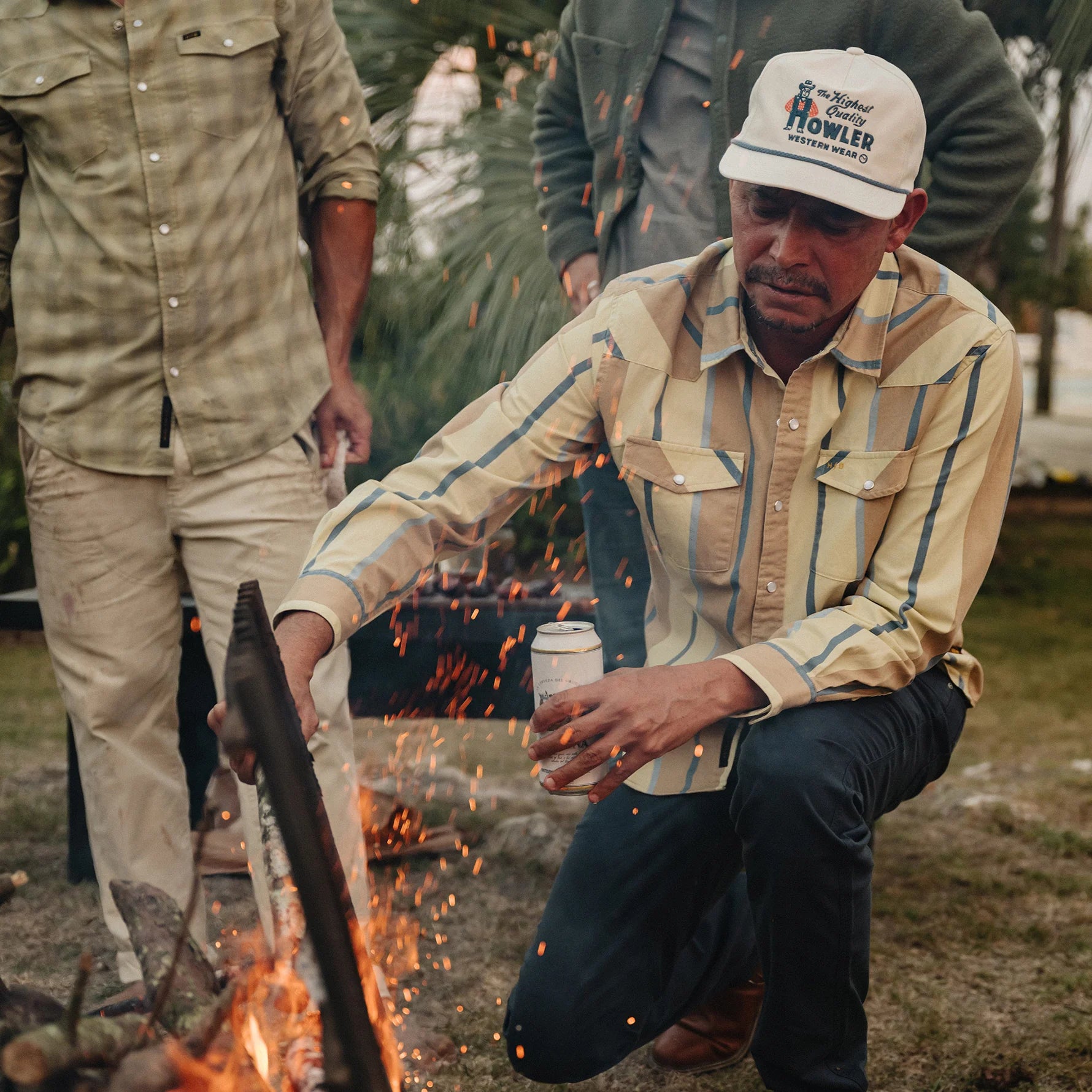 A man wearing a beige and blue striped button-up shirt, dark pants, and a white cap with 'Nimura Nowler' printed on it, crouches by a campfire outdoors while holding a cup. Sparks fly around him, and another person stands in the background.
