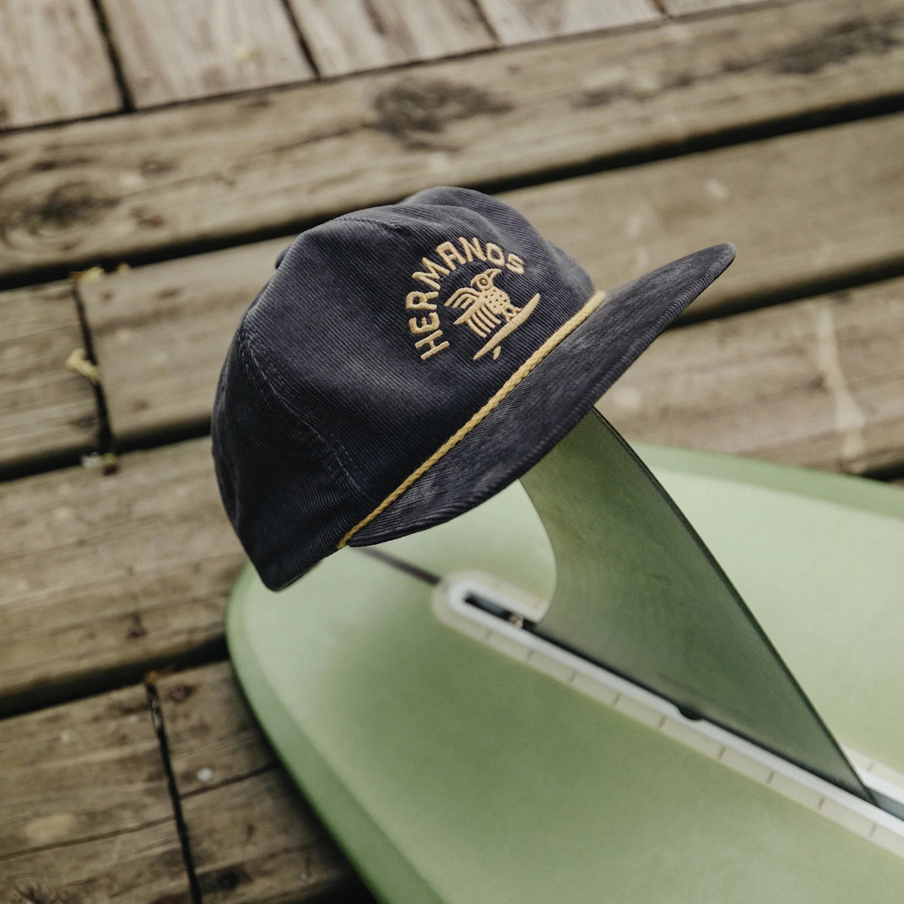 A dark navy blue corduroy cap with gold embroidered text reading 'HERMANDS' and a gold piping detail along the brim, resting on a light green surfboard over a rustic wooden deck.