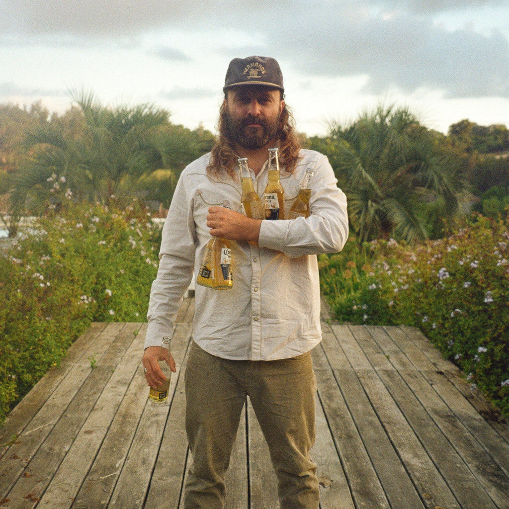 A man with a long beard and cap stands on a wooden boardwalk outdoors, holding multiple beer bottles against his chest. He is wearing a light-colored button-up shirt and khaki pants, with greenery and trees in the background under a cloudy sky.