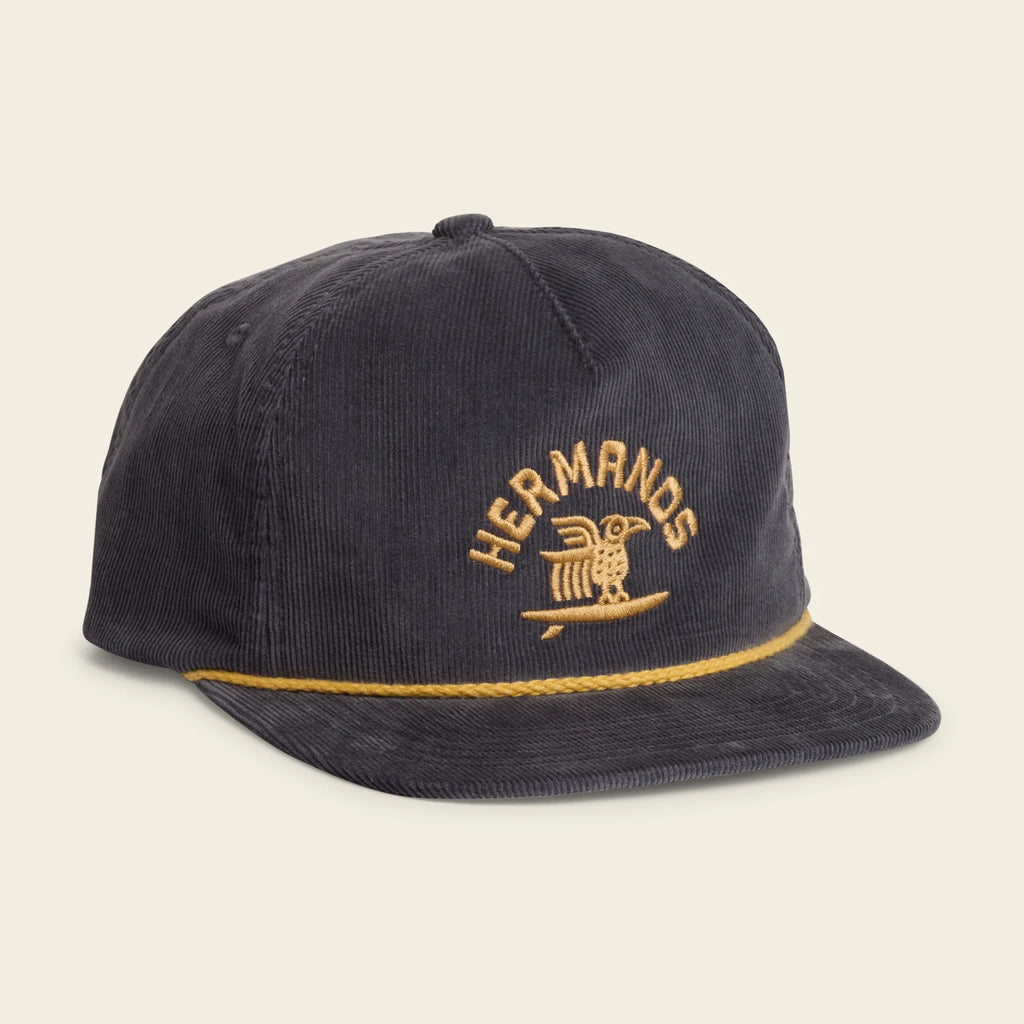 A dark navy corduroy baseball cap with gold embroidered 'HERMANDS' text and an owl logo on the front, plus a gold braided trim along the edge of the flat brim, shown on a light cream background.
