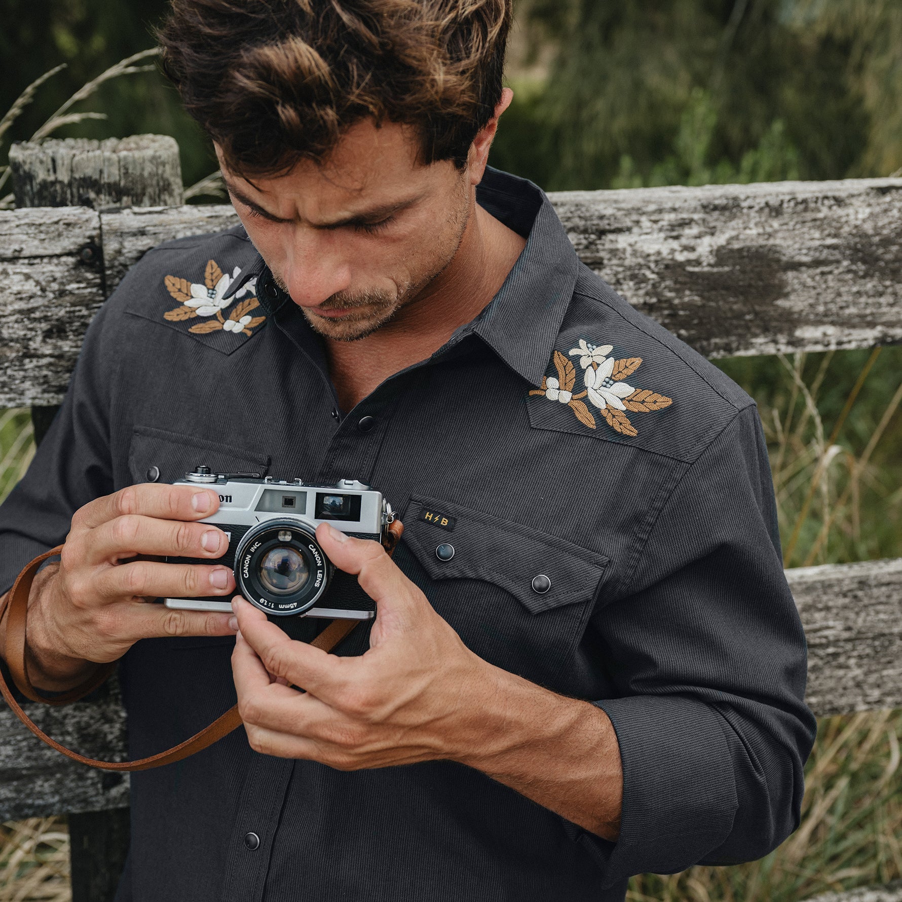A man wearing a dark charcoal gray western-style button-down shirt with embroidered white and gold floral designs on the shoulders, holding a vintage silver camera with a brown leather strap. He is posed outdoors in front of a weathered wooden fence with green foliage in the background.