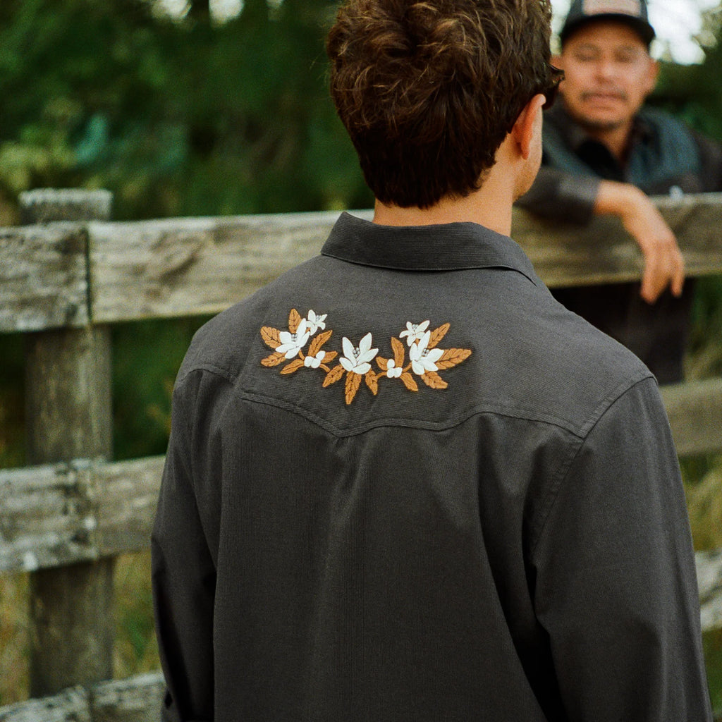 A man viewed from behind wearing a dark charcoal gray western-style shirt jacket with three embroidered floral motifs on the back shoulders. Another man is visible in the background behind a rustic wooden fence.