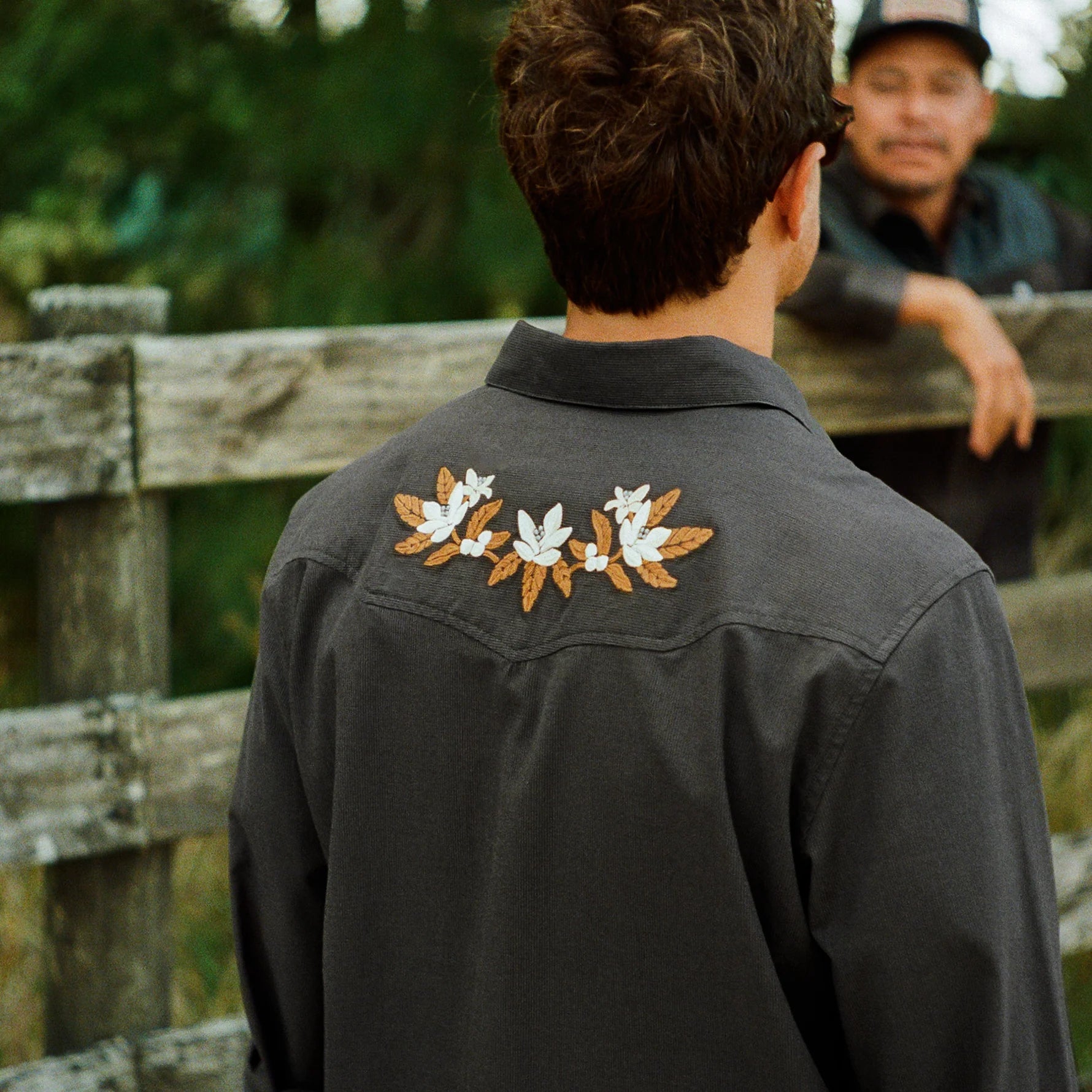 A man viewed from behind wearing a dark charcoal gray western-style shirt jacket with three embroidered floral motifs on the back shoulders. Another man is visible in the background behind a rustic wooden fence.