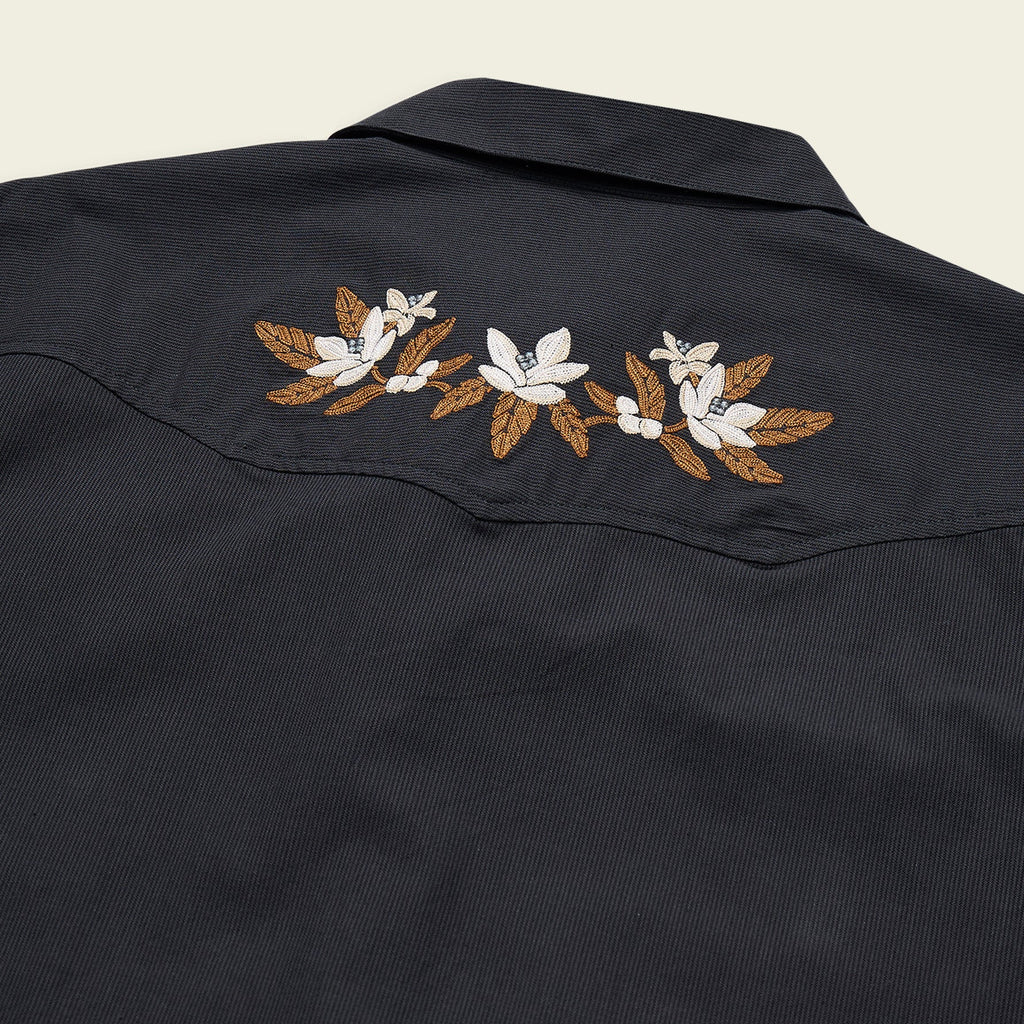 Close-up of a dark navy western-style shirt jacket showing embroidered white flowers and brown leaves across the shoulder yoke, set against a light cream background.
