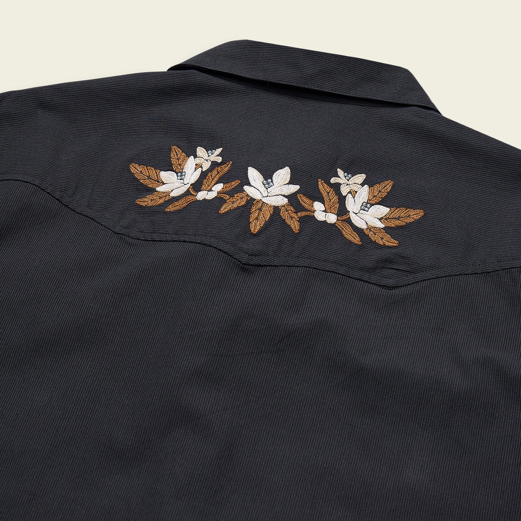 Close-up of a dark navy western-style shirt jacket showing embroidered white flowers and brown leaves across the shoulder yoke, set against a light cream background.