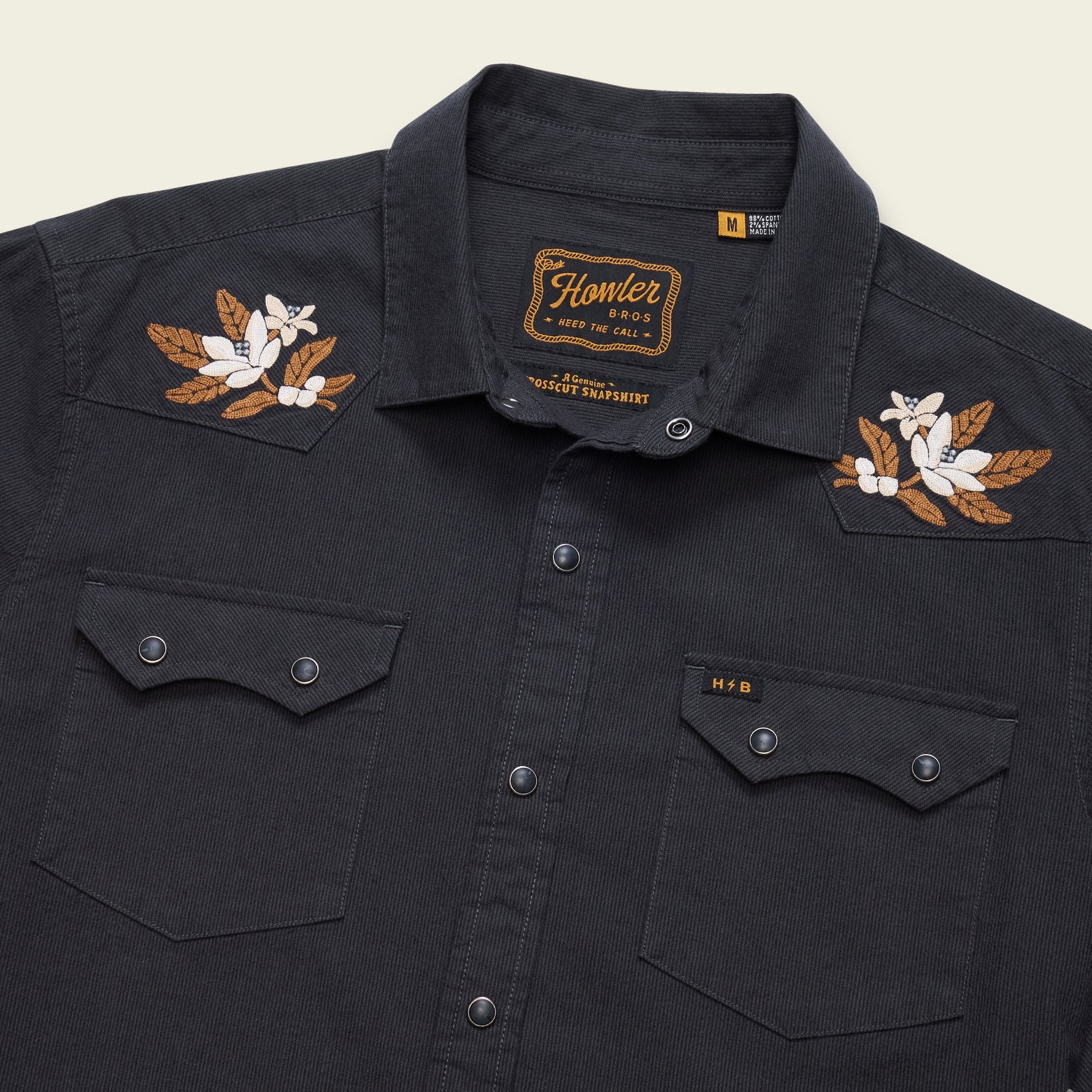 A close-up of a black western-style button-up shirt by Homer, featuring embroidered floral designs on the shoulders in brown and white, with two buttoned chest pockets.