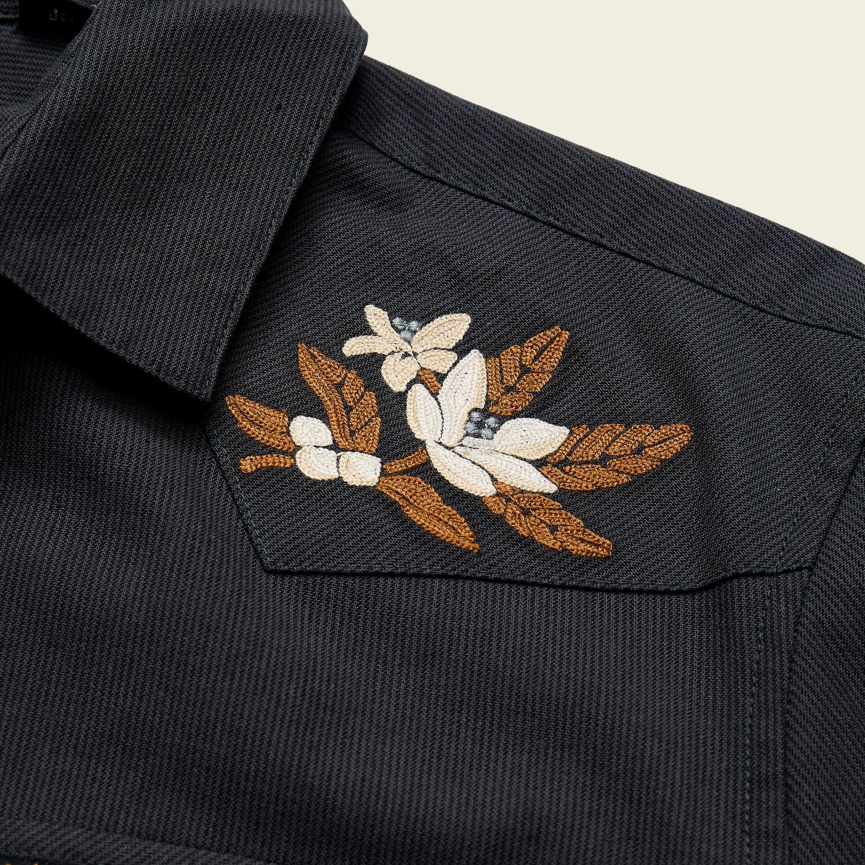 Close-up of a dark charcoal gray textured shirt jacket featuring a detailed floral embroidery on the chest pocket with white flowers, brown leaves, and small blue accents.