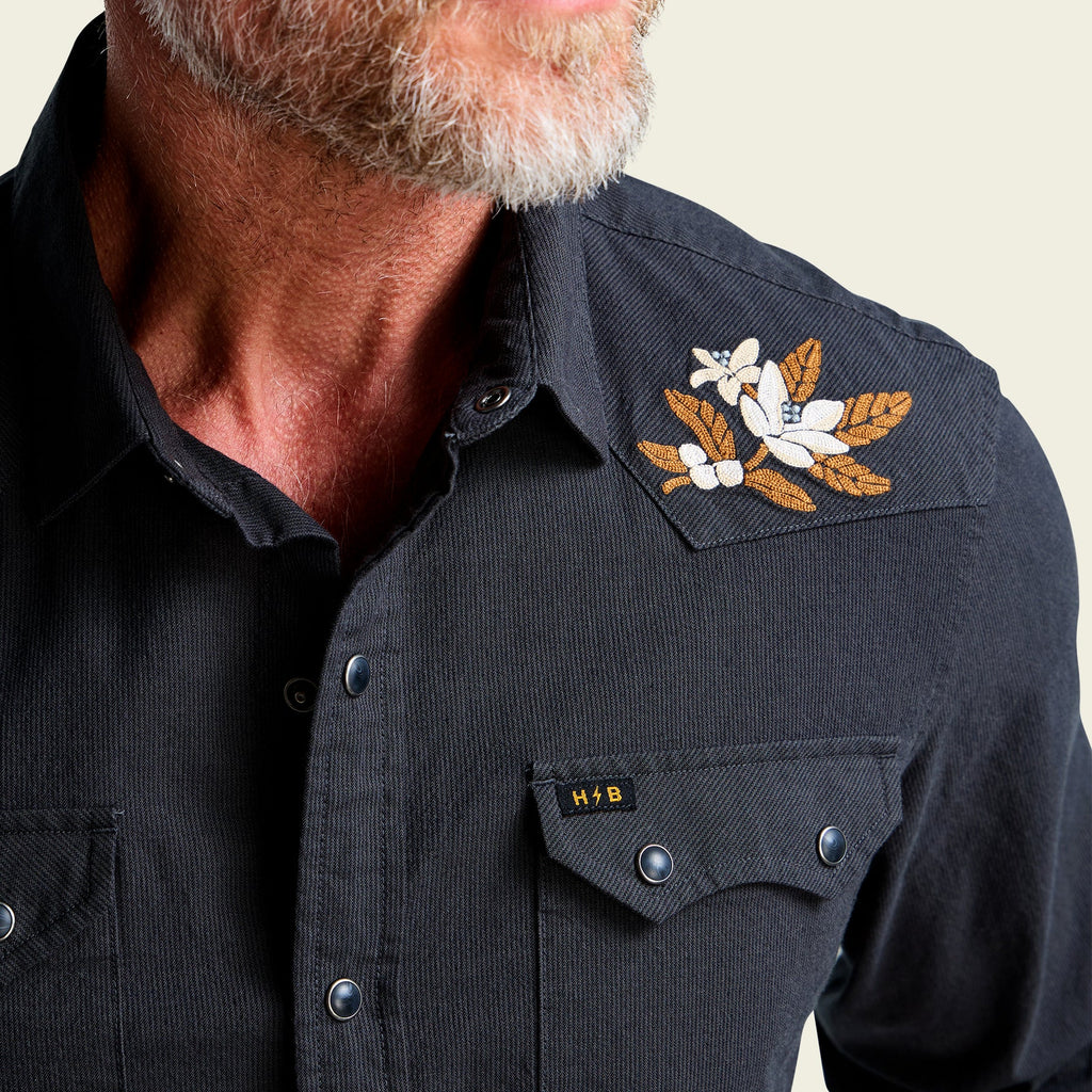 Close-up of a man wearing a dark charcoal corduroy western shirt with a floral embroidered patch on the shoulder and a chest pocket featuring a small HFB logo.