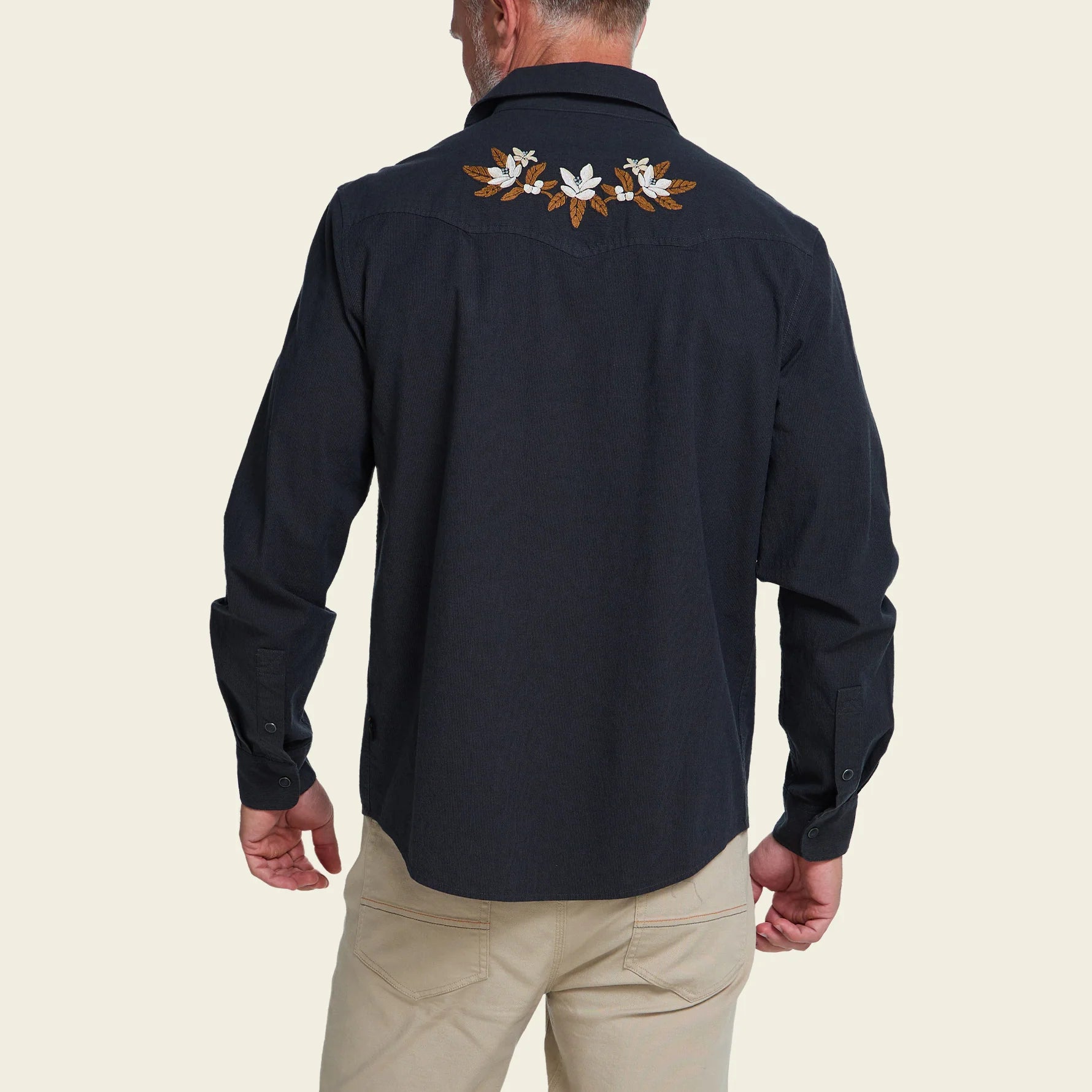 A man viewed from the back wearing a dark navy long-sleeve western-style shirt with white and brown floral embroidery across the upper back, paired with khaki pants, against a solid light beige background.