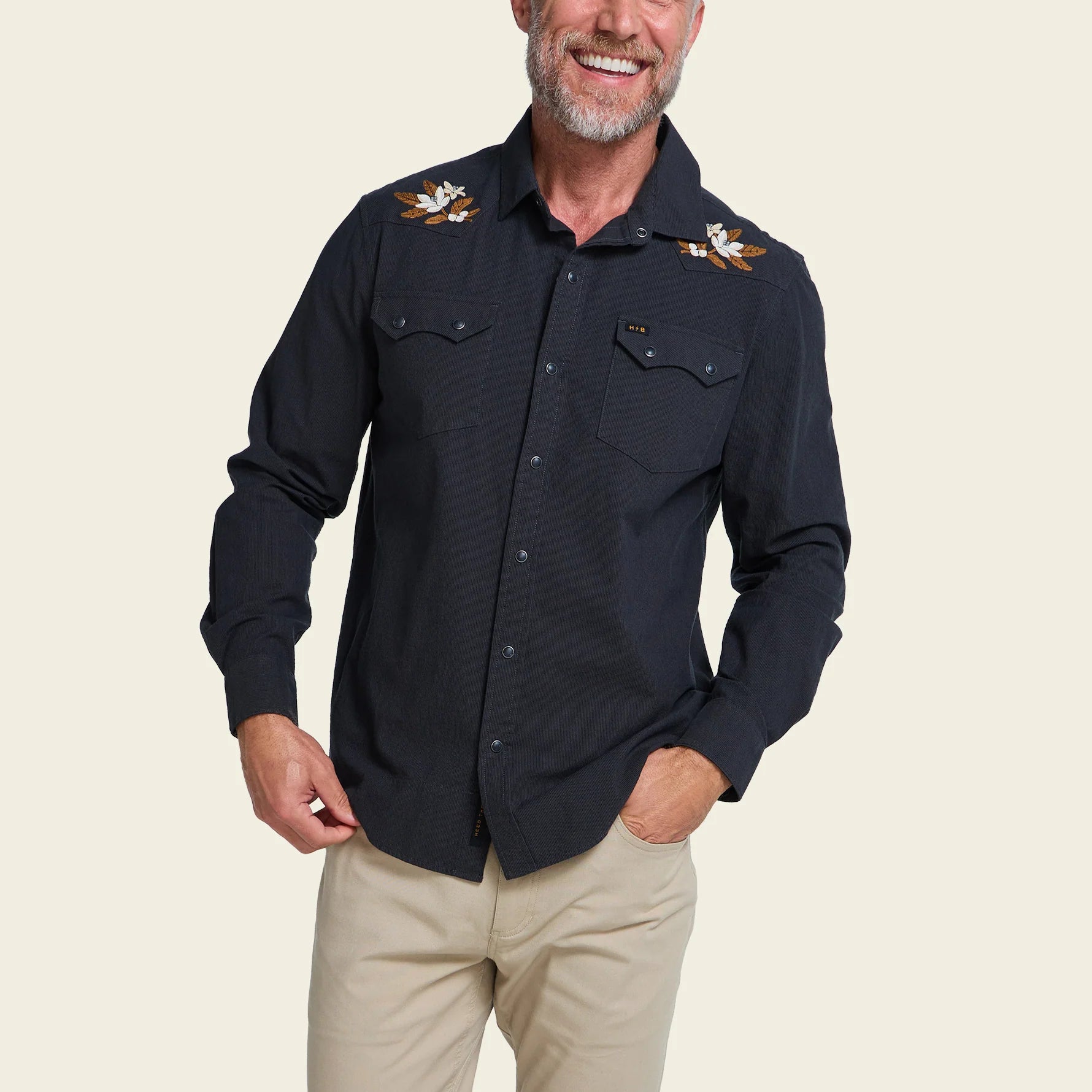 A smiling man wearing a dark navy western-style button-up shirt with embroidered floral details on the shoulders and beige pants, photographed against a plain light beige studio background.