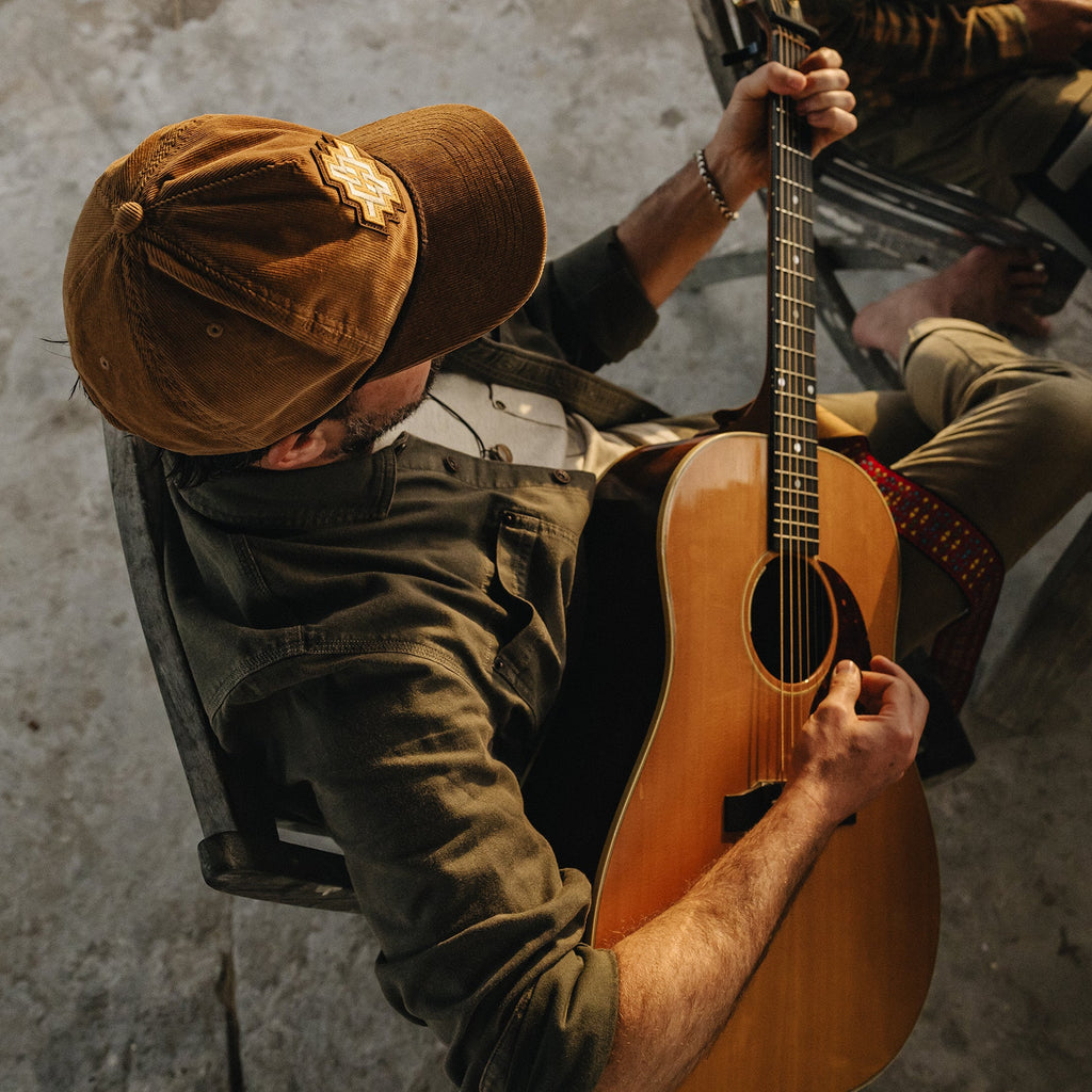 A man wearing a brown baseball cap and olive green shirt sits in a chair playing an acoustic guitar in a dimly lit indoor setting.